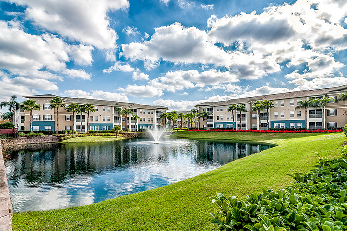 Charming apartment community with palm-lined pathways, tranquil pond, and elegant fountain under a vibrant blue sky.
