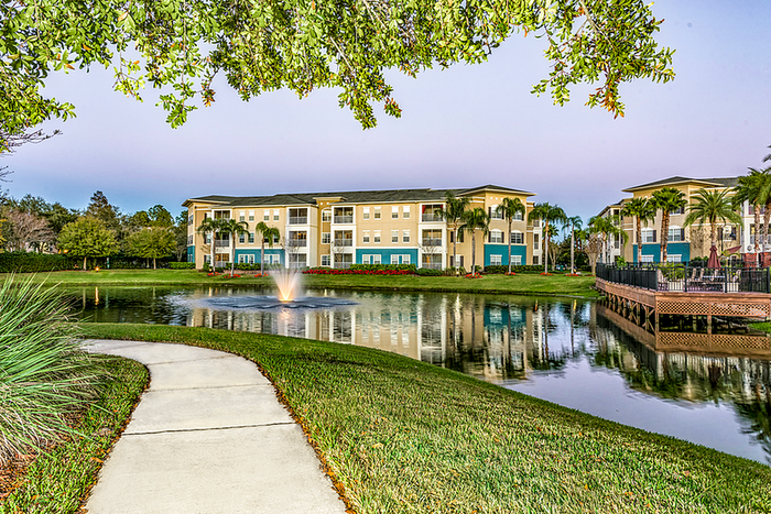 Scenic apartment community with palm trees, reflecting pond, and elegant fountain under a pastel evening sky.