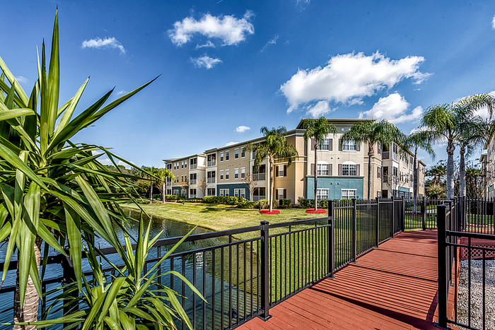 Elegant apartment community with modern architecture surrounded by lush palm trees and a serene pond under a bright blue sky.