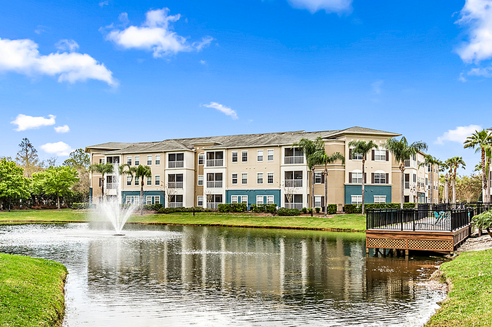 Charming apartment community with palm trees, overlooking a serene pond with a fountain and blue skies.