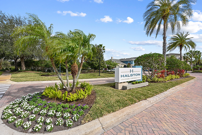 Inviting entrance of Halston Park Central with lush landscaping, palm trees, and beautiful pathways under a bright blue sky.