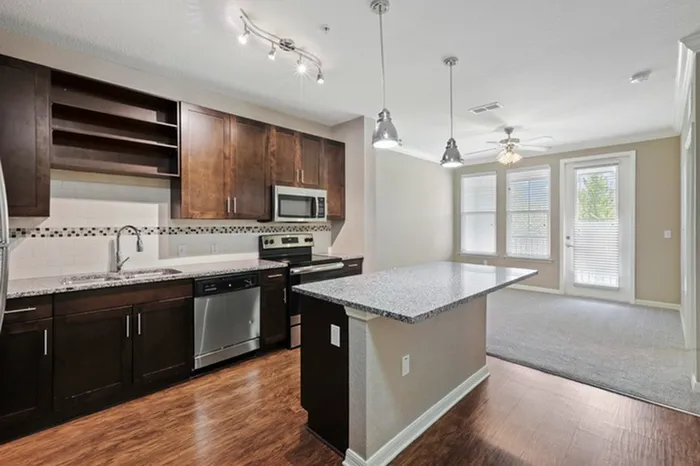 Kitchen with granite counters, wood plank floors, espresso custom cabinetry, stainless steel appliances, and a living room in the background with two large windows and a door leading to the balcony at Lantower Asturia.