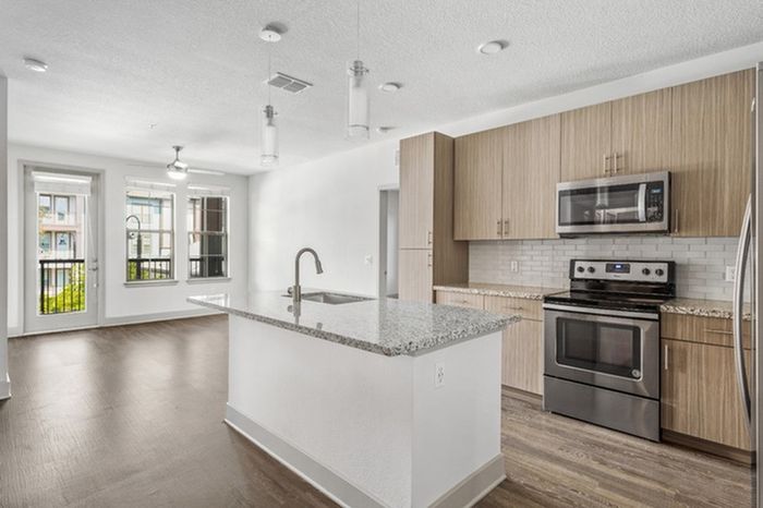 An apartment's energy-efficient kitchen with a side-by-side refrigerator, granite countertops, stainless steel appliances, and a kitchen island with the hard-wood floor living room the background with access to a balcony at Lantower Grande Flats Apartments.