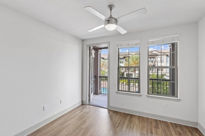 Living room space with a ceiling fan, hardwood floors, two large windows, and a door leading to a balcony facing the resort-style pool and palm trees at Lantower Grande Flats.