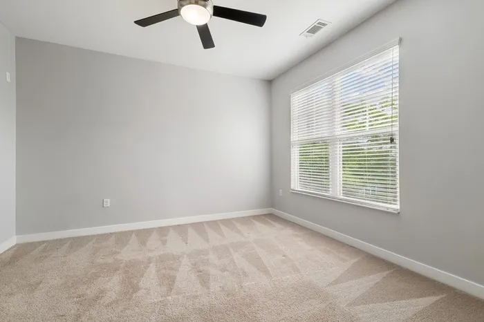 Carpeted bedroom with a ceiling fan and large window with natural sunlight at Realm at Patterson Place.
