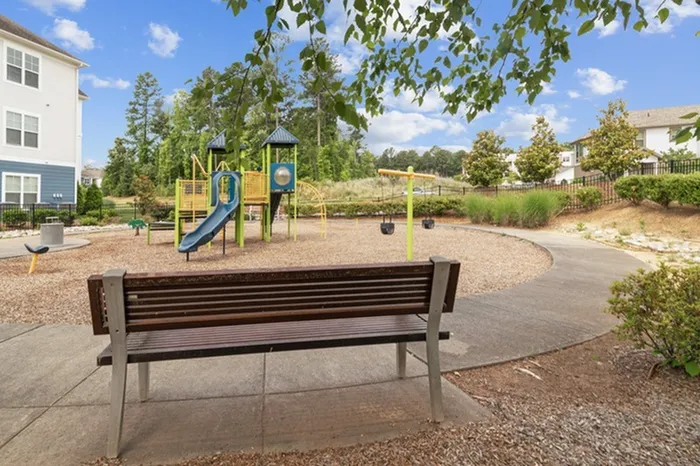 Fenced playground with wood chips, swings, a play structure, and bench at Patterson Place Apartments.