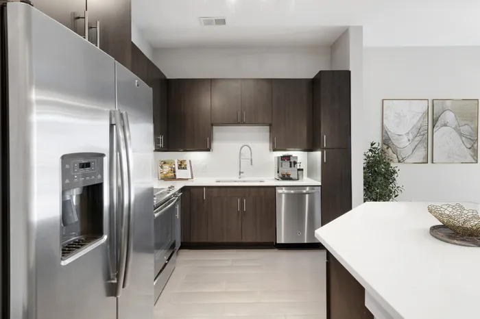 Large, open concept kitchen at Bullhouse with wood-style flooring, stainless steel, energy efficient appliances, white quarts countertops, a kitchen bar, and tile backsplash decorated with a coffee machine, house plant, decorative bowl and grey wall art.