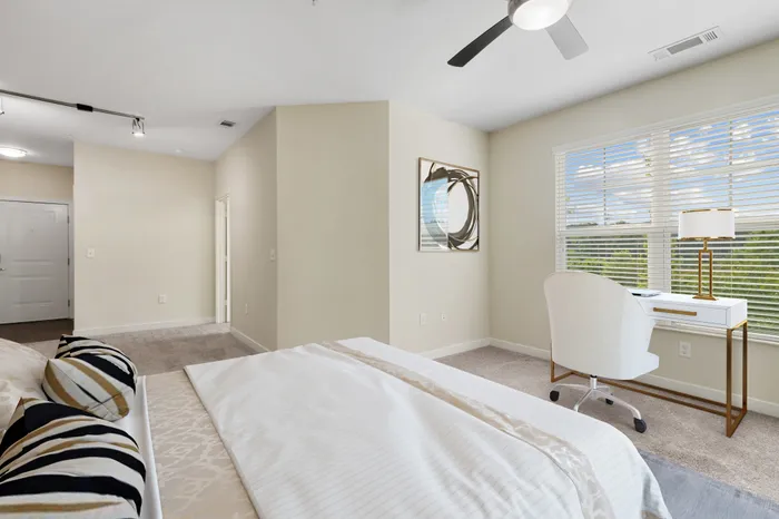 Bedroom with tan carpet, a ceiling fan, 9-foot ceilings, large windows, a large, cream bed and modern wall art at Realm at Patterson Place Apartments.