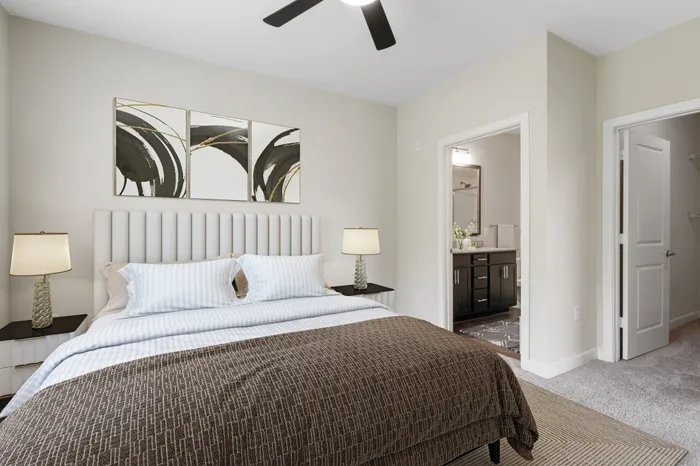 Bedroom with tan carpet, a ceiling fan, 9-foot ceilings, large windows, a large, cream bed with brown bedding and modern wall art at Realm at Patterson Place Apartments.