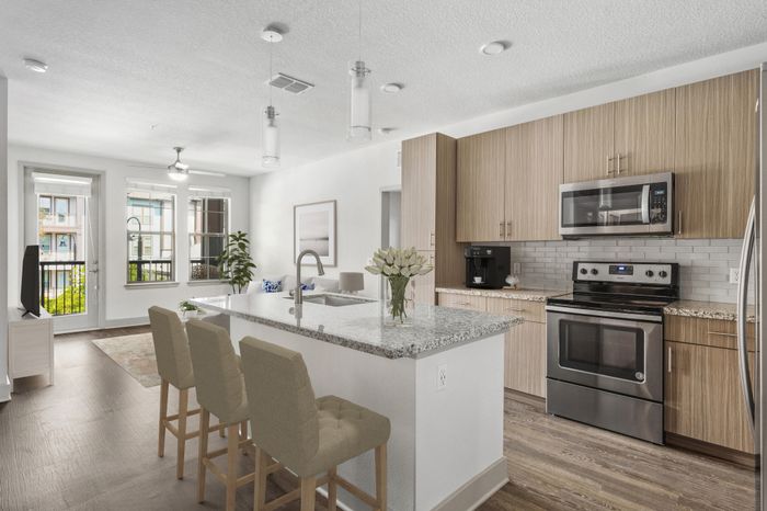 An apartment's energy-efficient kitchen with a side-by-side refrigerator, granite countertops, stainless steel appliances, and a kitchen island with hard-wood flooring with access to a private balcony at Lantower Grande Flats Apartments decorated with modern kitchen stools and a bouquet of flowers.