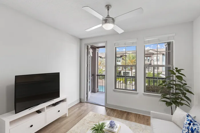 Living room space with a cream couch, blue decorations, a TV with a white stand, a houseplant, a ceiling fan, hardwood floors, 10-foot ceilings, and access to a private balcony facing the resort-style pool and palm trees at Lantower Grande Flats.