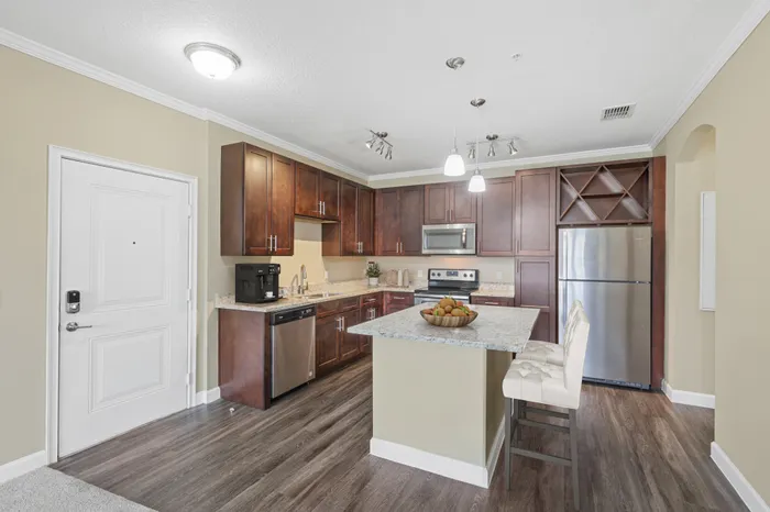 A kitchen with espresso wood cabinetry, wood-style flooring, a kitchen island with a granite countertop, Energy Star Stainless Steel Appliances, and a built-in wine soffit at Lantower Grande Pines decorated with a fruit bowl and coffee machine.