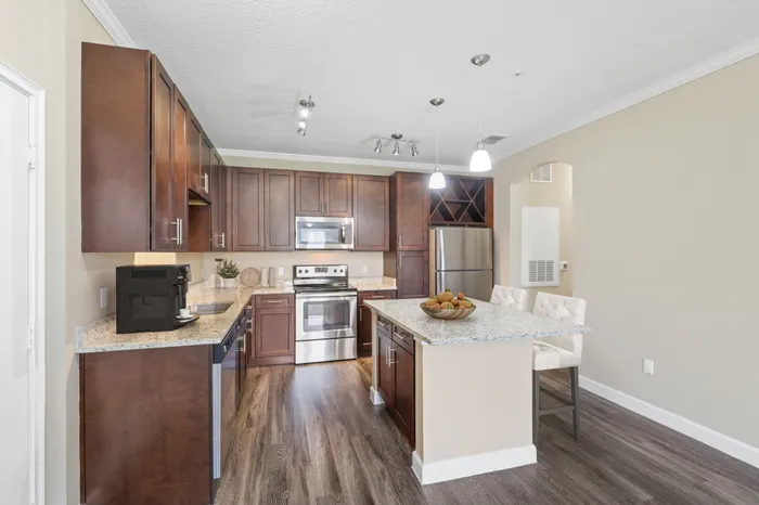 A kitchen with espresso wood cabinetry, wood-style flooring, a kitchen island with a granite countertop, Energy Star Stainless Steel Appliances, and a built-in wine soffit at Lantower Grande Pines decorated with a fruit bowl, stools and a coffee machine.