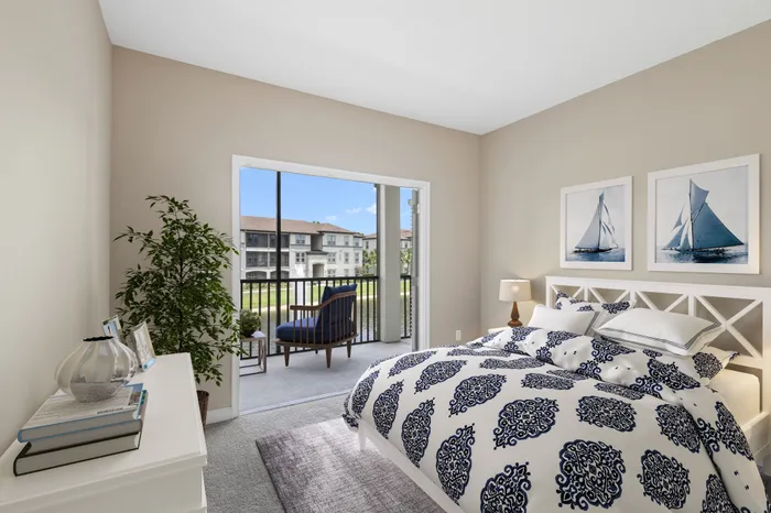 A carpeted room with a large white bed and dresser with blue decorations, a tall plant and open French doors leading to the balcony with a view of a pond with a fountain and trees at Lantower Grande Pines.