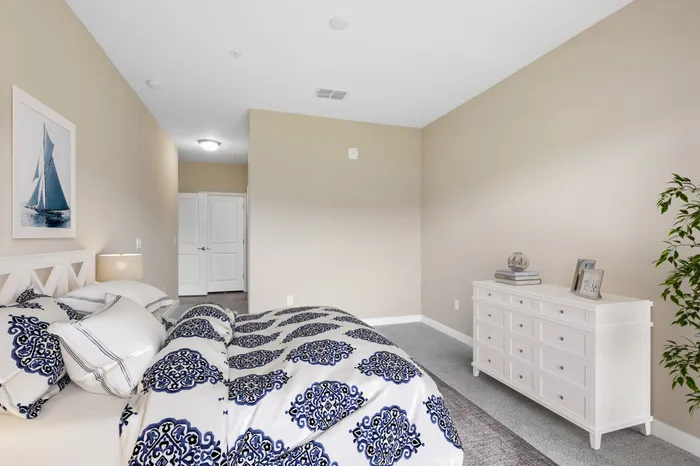 A carpeted, spacious bedroom with cream walls, a large white bed, a white dresser, blue decorations and a tall plant at Lantower Grande Pines.