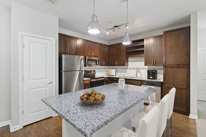 Kitchen with Luna Pearl granite countertops, wood-style plank floors, espresso custom cabinetry, a stainless-steel whirlpool appliance package, and tile backsplash with white bar stools, a bowl of fruit and a coffee machine at Lantower Asturia.