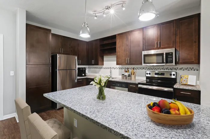 Kitchen with Luna Pearl granite countertops, wood-style plank floors, espresso custom cabinetry, a stainless-steel whirlpool appliance package, and tile backsplash with white bar stools, a bowl of fruit and a coffee machine at Lantower Asturia