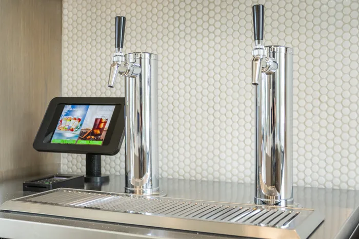 Modern beverage dispensing system with digital touchscreen interface and dual chrome taps, set against elegant tile backsplash.