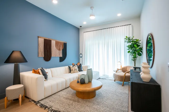 Modern living room with elegant decor, featuring a cozy white sectional sofa, stylish wooden coffee table, and vibrant blue accent wall adorned with artistic hanging. Natural light pours through sheer curtains, illuminating the space with warmth and charm.
