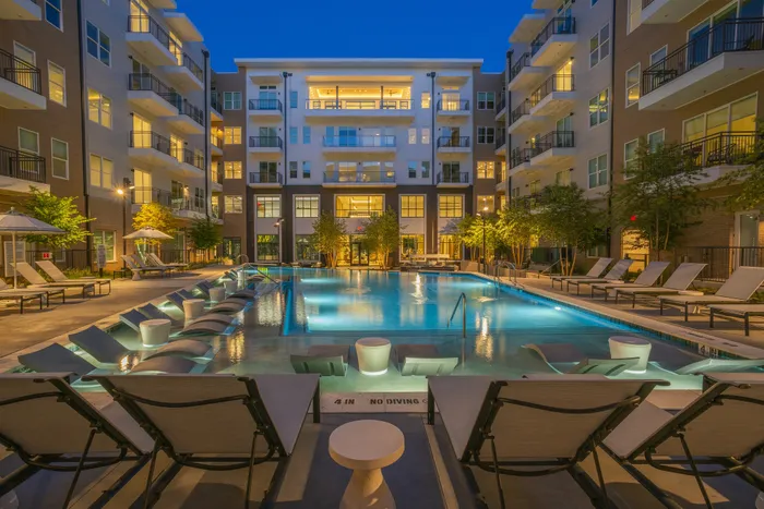 Luxurious apartment complex with a beautiful illuminated swimming pool, surrounded by modern balconies and stylish lounge chairs, creating a serene evening ambiance.