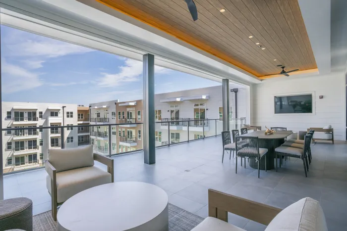 Luxurious covered balcony with modern seating and dining area, overlooking scenic apartment architecture and bright blue skies.