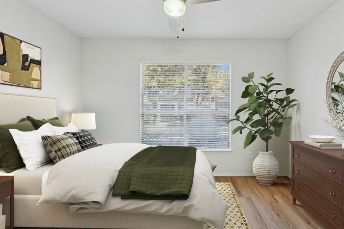Inviting bedroom with stylish decor, featuring a cozy bed with accent pillows, large window with blinds, and a lush potted plant beside a wooden dresser.