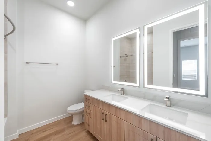 Modern bathroom with elegant double vanity, illuminated mirrors, and sleek wooden cabinetry against a bright, minimalist backdrop.