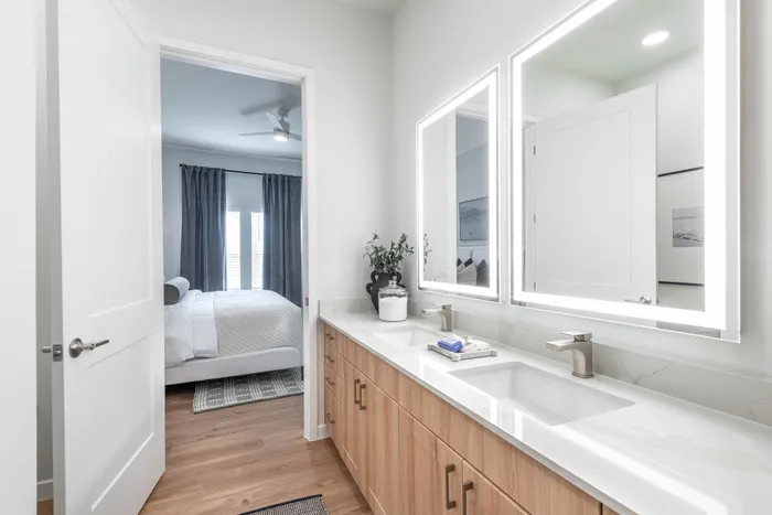 Elegant bathroom with dual sinks and stylish vanity, leading to a cozy bedroom with natural light and modern decor.