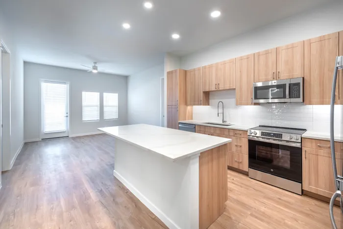 Modern kitchen with sleek wooden cabinetry, stainless steel appliances, and a spacious light quartz island, leading to a bright living area with large windows and elegant hardwood floors.