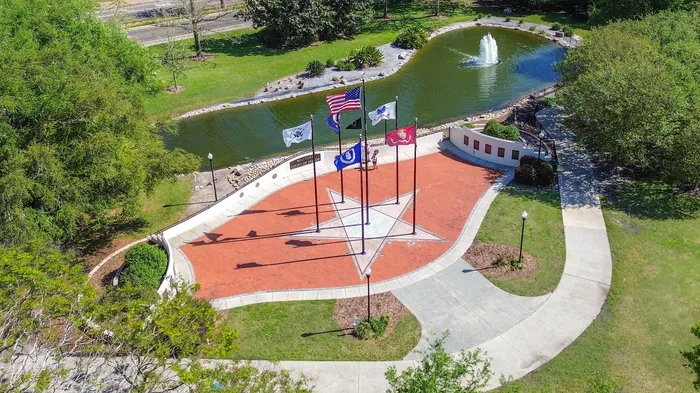 Aerial view of a serene park with a star-shaped plaza, multiple flags, and a tranquil pond featuring a fountain, surrounded by lush greenery.
