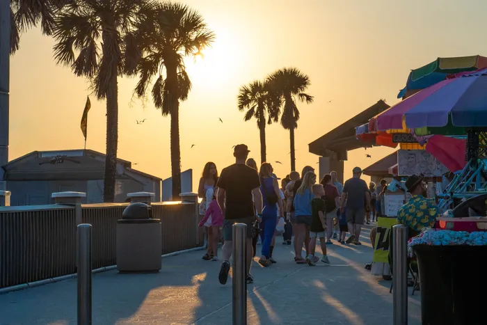 Vibrant boardwalk scene at sunset with palm trees, people strolling, colorful stalls, and a warm golden sky.