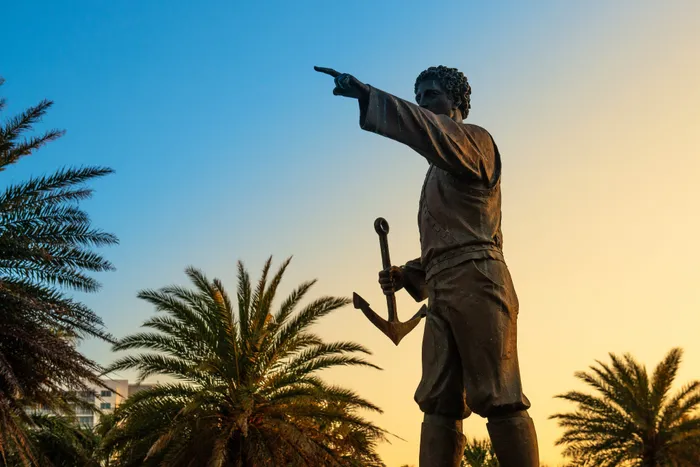 Majestic bronze statue of a sailor holding an anchor, set against a vibrant sunset sky and surrounded by palm trees.