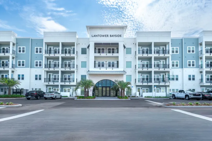 Modern four-story apartment building with spacious balconies and elegant entryway, surrounded by palm trees and ample parking.
