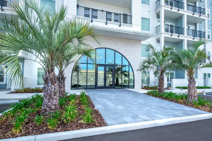 Contemporary apartment entrance with grand arched glass doors, surrounded by lush palm trees and elegant landscaping.