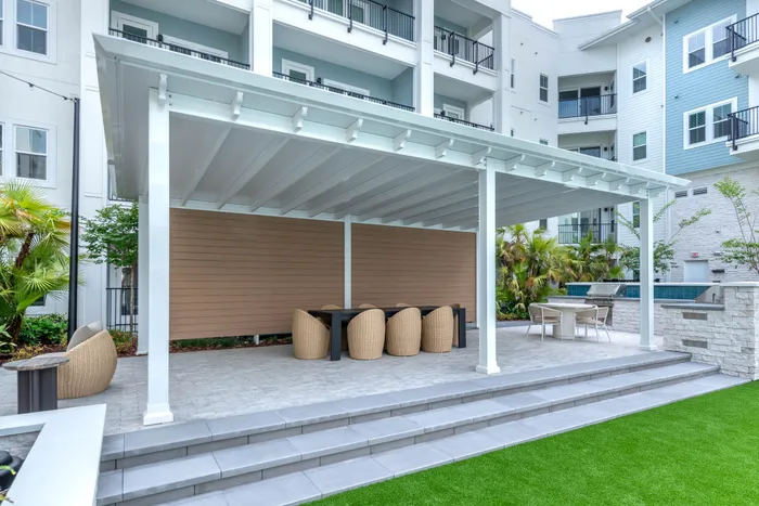 Elegant outdoor seating area with modern wicker chairs under a stylish pergola, featuring lush greenery and vibrant lawn in a luxury apartment complex.