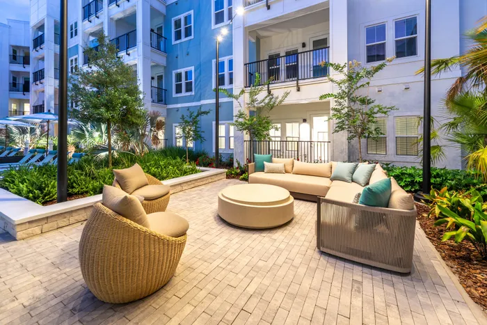 Chic outdoor lounge area with modern wicker seating and plush cushions, surrounded by lush greenery in a vibrant apartment complex courtyard.