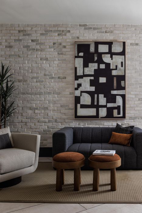 Elegant living room with modern decor featuring abstract art, plush sofas, and chic stools against a textured brick wall.