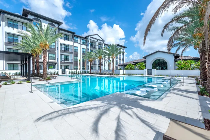 Luxurious apartment complex with a stunning outdoor swimming pool, surrounded by palm trees and modern architecture under a bright blue sky.