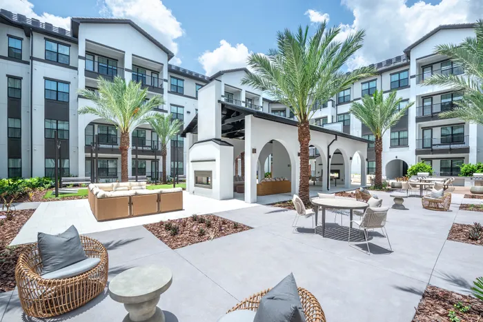 Modern apartment complex with elegant courtyard featuring palm trees, outdoor seating, and open-air lounge area under sunny skies.