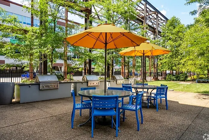 Charming outdoor patio area with vibrant blue chairs, sunny yellow umbrellas, and modern grills set amidst lush greenery.