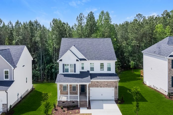Charming two-story home with a welcoming front porch, set against a lush wooded backdrop and featuring a well-maintained lawn and modern design.