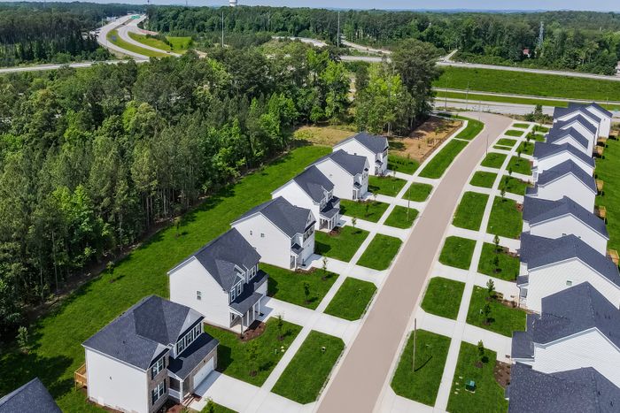 Aerial view of a picturesque suburban neighborhood featuring rows of contemporary houses with manicured lawns, set against a lush green forest backdrop and winding roadways nearby.