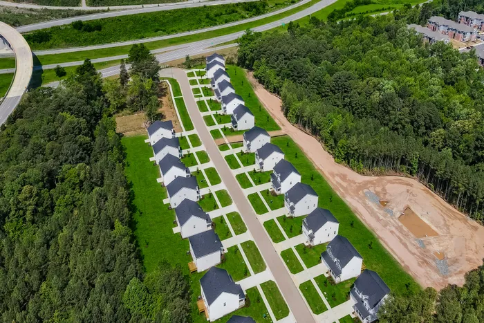 Aerial view of a picturesque housing development featuring modern single-family homes with manicured lawns, surrounded by lush greenery and easy highway access.