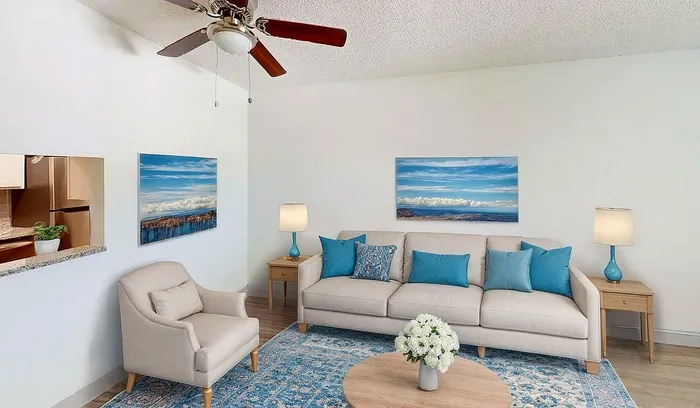 Charming living room with a cozy beige sofa, vibrant blue cushions, elegant armchair, and modern ceiling fan. Features a stylish open concept design with a glimpse into a contemporary kitchen.