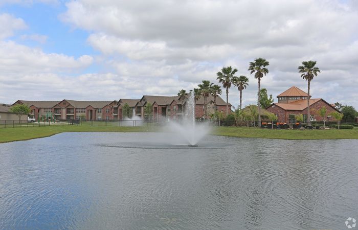 Serene lakeside view of modern brick apartments with palm trees and a central fountain under a blue sky.