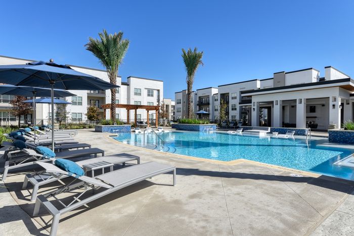 Luxurious apartment complex featuring a pristine outdoor pool with stylish lounge chairs and umbrellas, surrounded by modern architecture and palm trees.