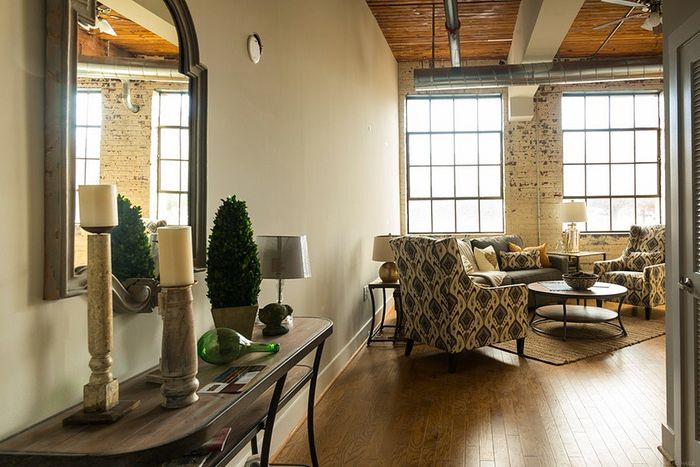 Chic living room in an industrial-style apartment, featuring large factory windows, cozy seating with patterned chairs, and elegant decor on wooden flooring.