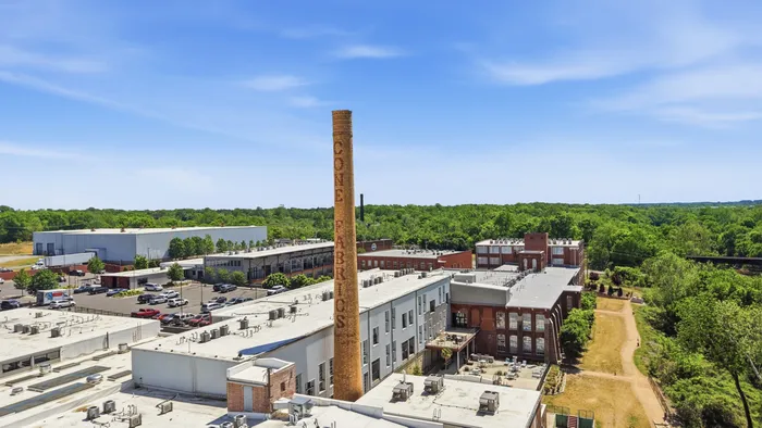 Urban residential area showcasing a historic brick smokestack labeled 'Cone Fabrics', surrounded by lush greenery and modern industrial architecture.