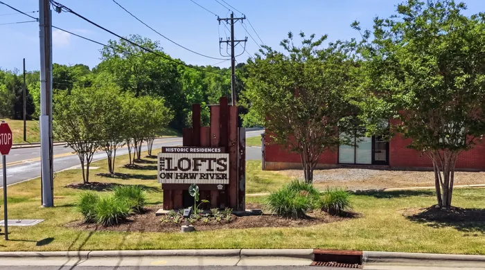 Inviting entrance to 'The Lofts on Haw River' featuring lush green landscaping and historic charm.