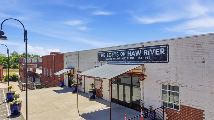 Charming historic building of 'The Lofts on Haw River' with red brick and white facade, featuring industrial-style architecture and vibrant blue planters under a clear blue sky.